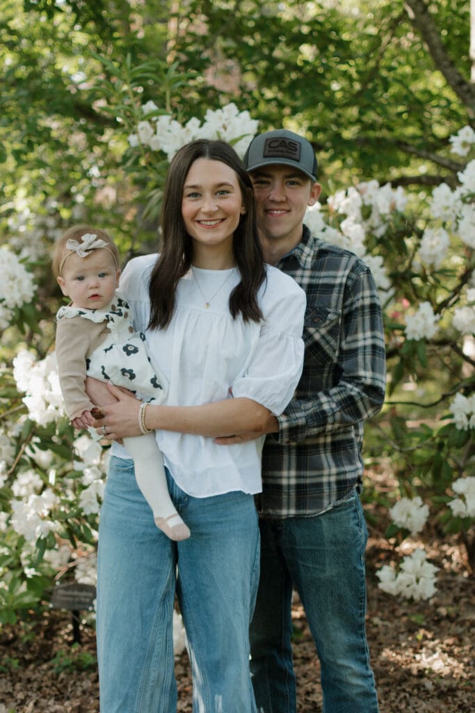 A family of 3 standing in front of some flowers and smiling