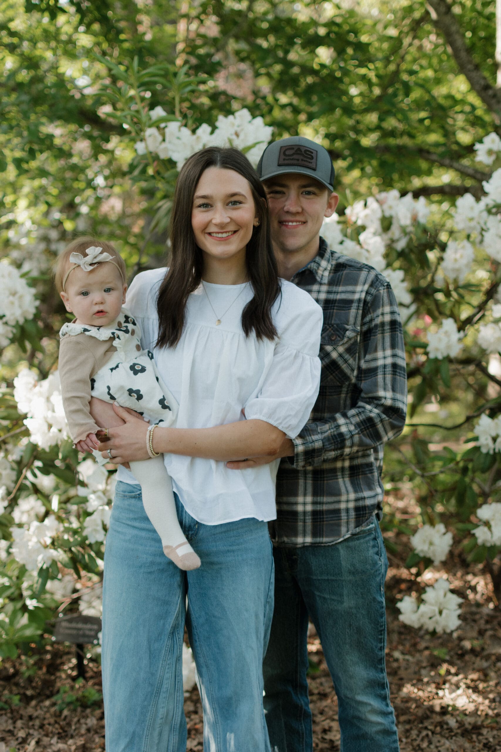A family of 3 standing in front of some flowers and smiling