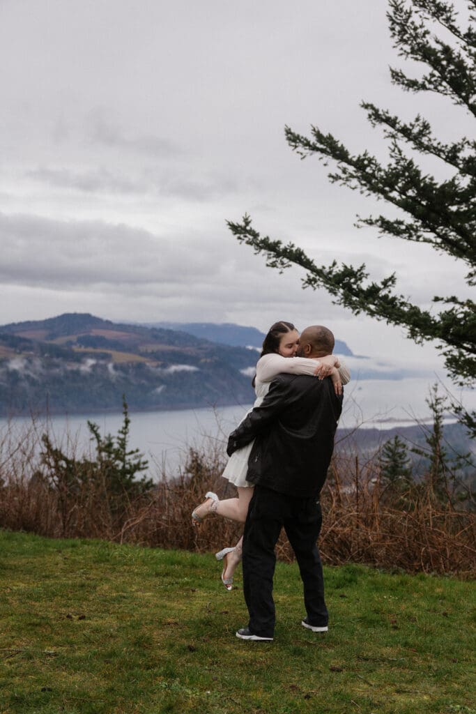 Partner lifting fiancé on grassy cliffside at Portland Women’s Forum State Scenic Viewpoint in Corbett, Oregon.