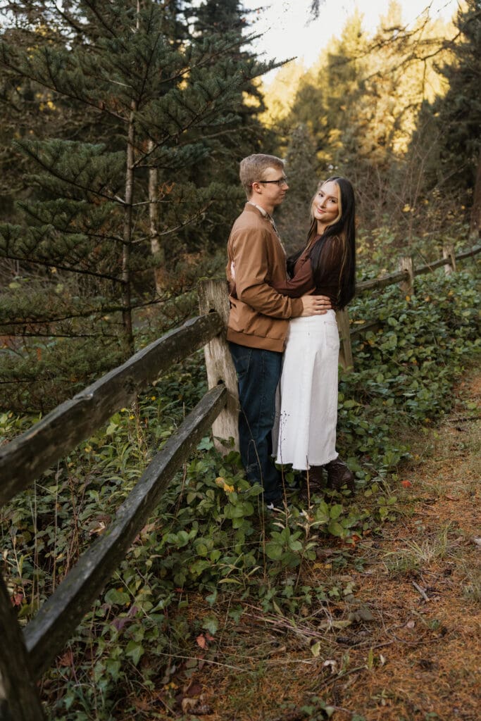 Couple embracing beside a wooden fence surrounded by evergreen trees at Hoyt Arboretum Portland Oregon fall engagement session