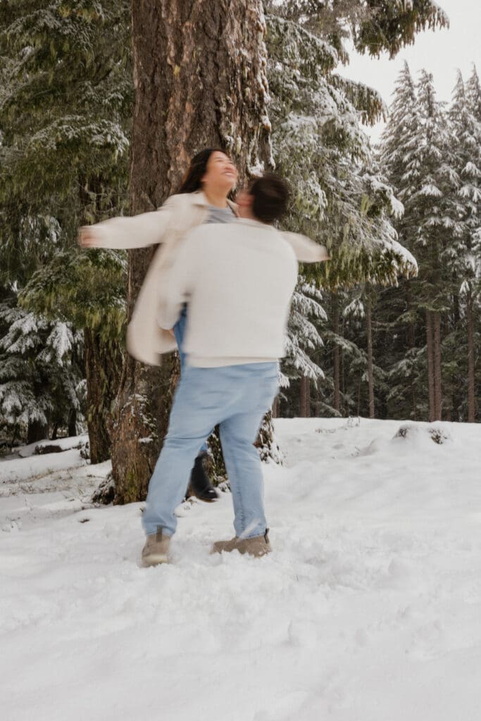 Couple spinning in the snow beneath pine trees during a Mount Hood engagement session in Oregon