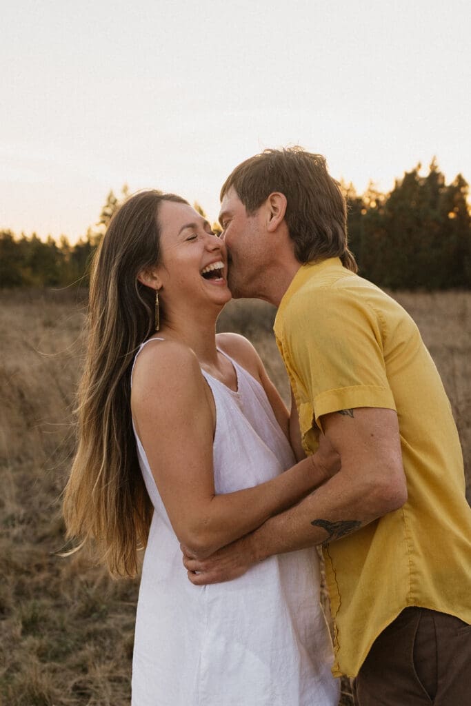 Engaged couple laughing together in a golden field at Graham Oaks Nature Park in Wilsonville, Oregon during sunset engagement session