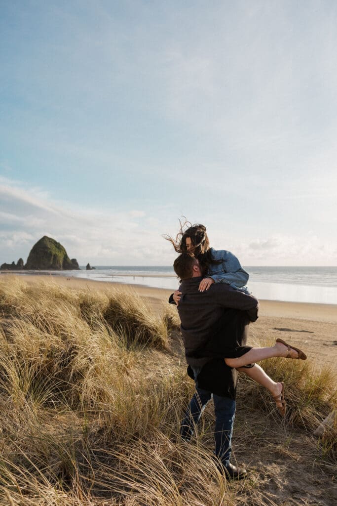 Man lifting his partner in the sand dunes at Cannon Beach Oregon engagement session with Haystack Rock in the background