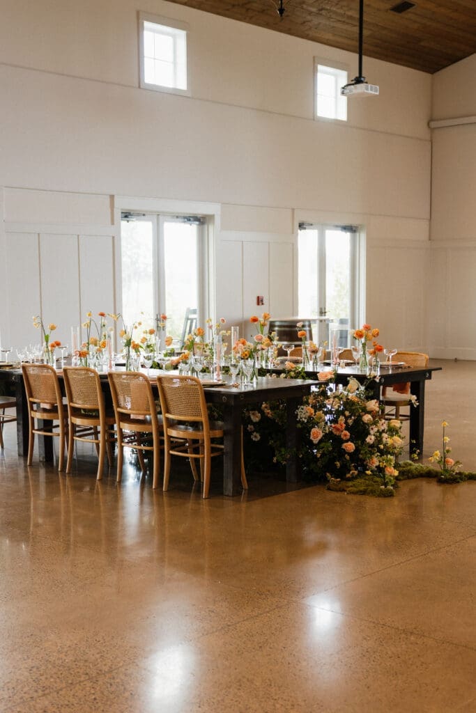 Long reception table styled with orange and peach florals inside the bright modern farmhouse event center at The Farm on Golden Hill in Silverton Oregon
