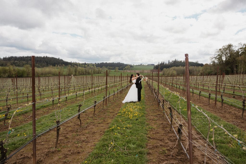 Bride and groom kissing between vineyard rows at The Farm on Golden Hill with rolling Oregon hills in the background