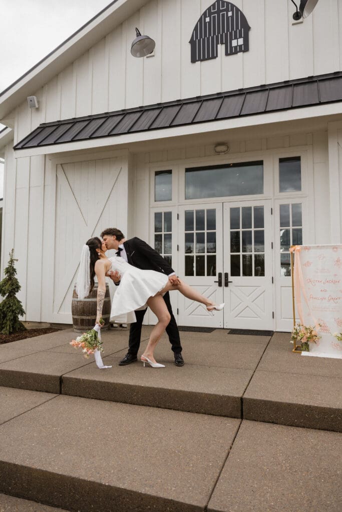 Groom dipping bride in a kiss in front of the white barn doors of The Farm on Golden Hill event center in Silverton Oregon