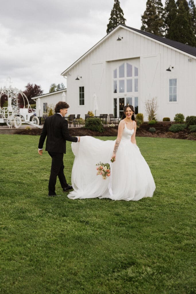 Bride and groom walking across the lawn at The Farm on Golden Hill wedding venue in Oregon, bride laughing as groom carries her train