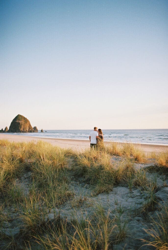 Couple standing in coastal dunes during a film maternity session at Cannon Beach, Oregon, with Haystack Rock in the background.