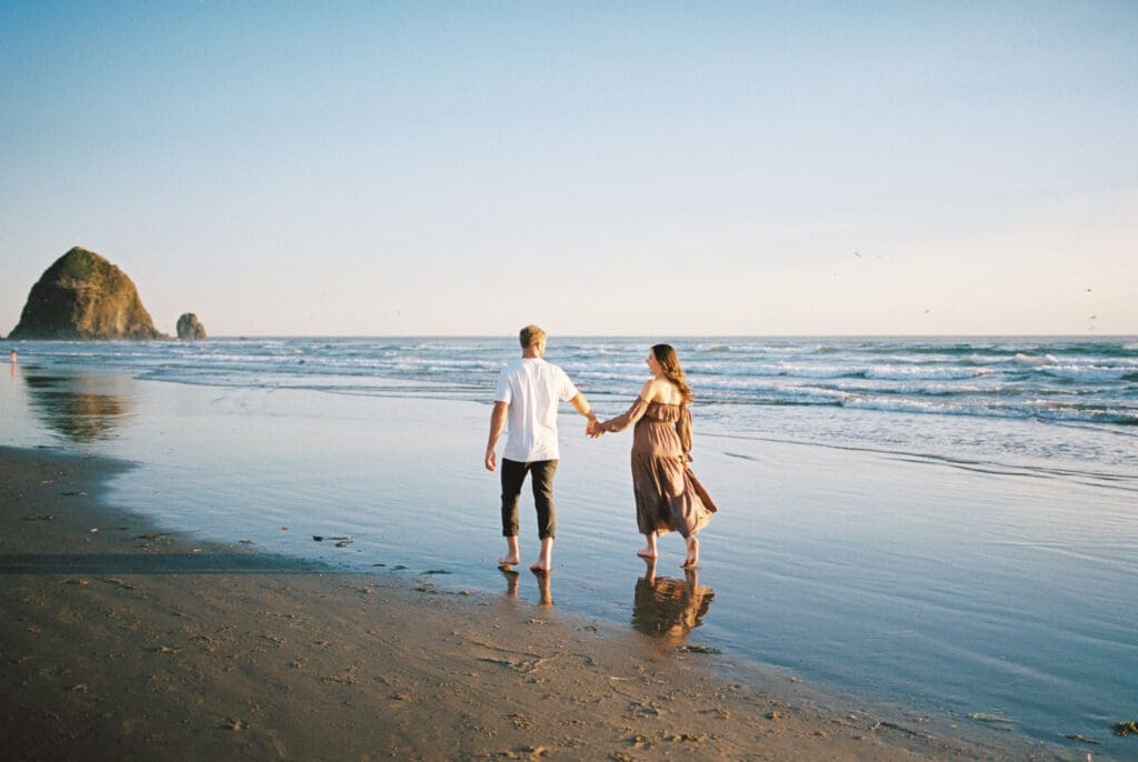 Couple holding hands while walking along the shoreline during a film maternity session at Cannon Beach, Oregon.