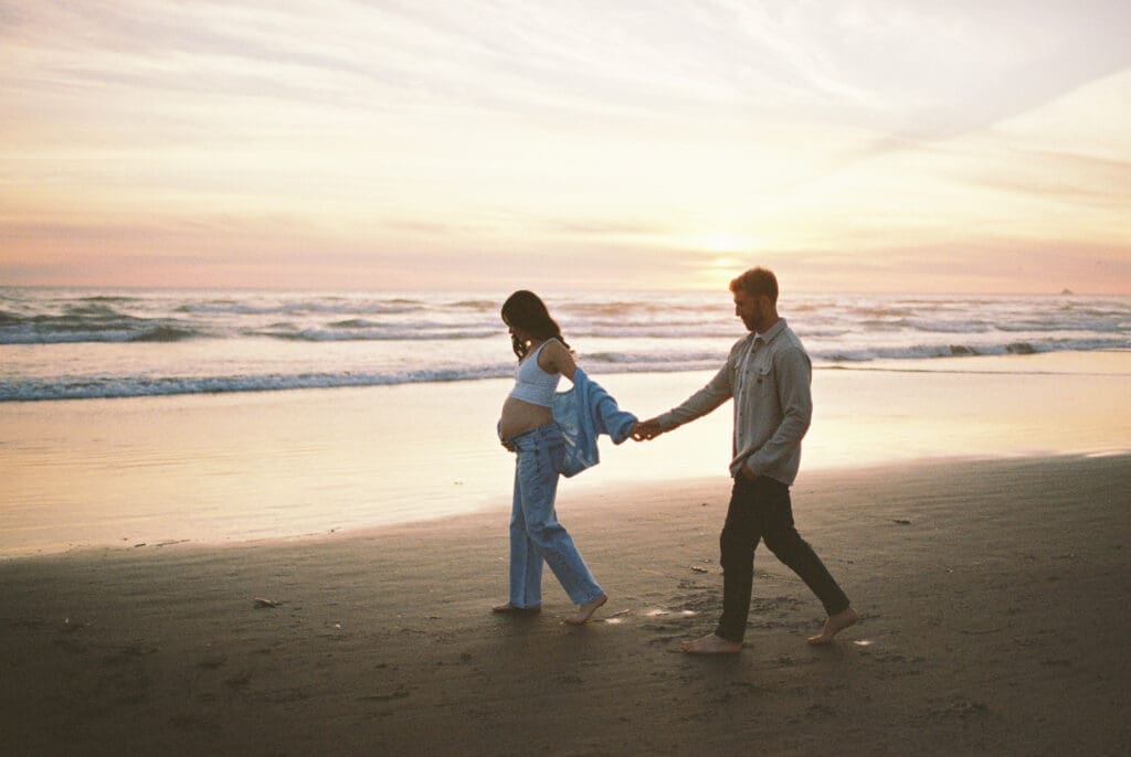 Pregnant person and partner walking hand in hand at sunset during a film maternity session at Cannon Beach, Oregon.
