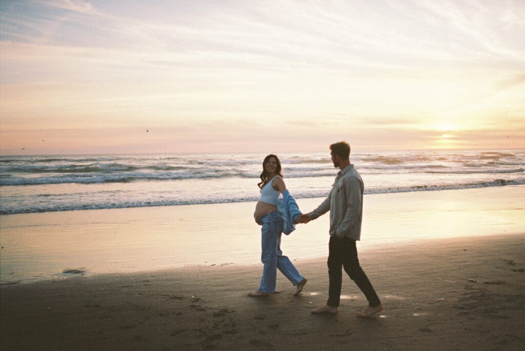 Pregnant person and partner walking hand in hand at sunset during a film maternity session at Cannon Beach, Oregon.