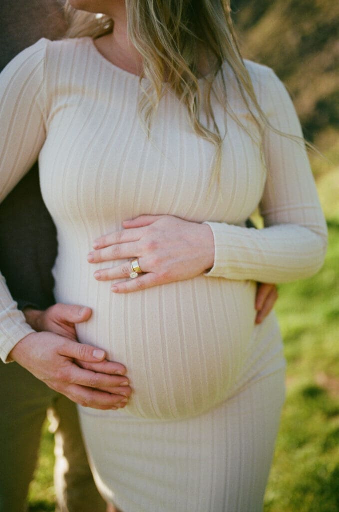 Partner embracing a pregnant person and holding their baby bump during a film maternity session at Cannon Beach, Oregon.